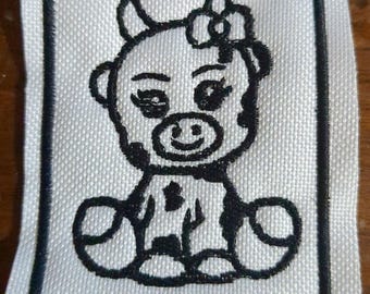May include: A square white fabric patch featuring a black embroidered outline of a seated cow character. The cow has spots, a small bow on its head, and is enclosed within a thick black rectangular border stitched onto the white fabric background.