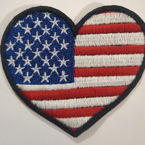 May include: A heart-shaped patch with an embroidered American flag design. The flag is red, white, and blue with 13 stripes and 50 stars.