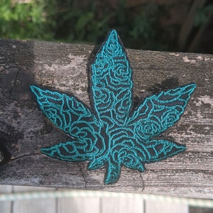Könnte beinhalten: Ein gestickter Patch in blaugrüner Farbe, der die Form eines Marihuana-Blattes mit einem wirbelnden Muster hat.