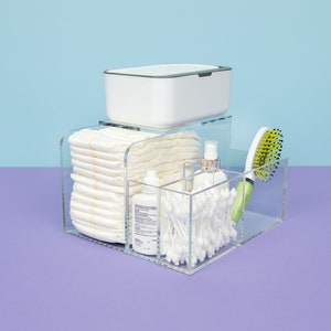 May include: A clear acrylic organizer with compartments for baby supplies. The organizer holds a stack of diapers, a bottle of lotion, a container of cotton swabs, and a hairbrush. The organizer is on a purple surface.
