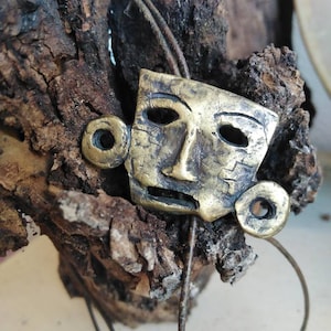 May include: A gold-toned metal pendant in the shape of a face with stylized features. The pendant is hanging from a brown cord and is resting on a piece of weathered wood.