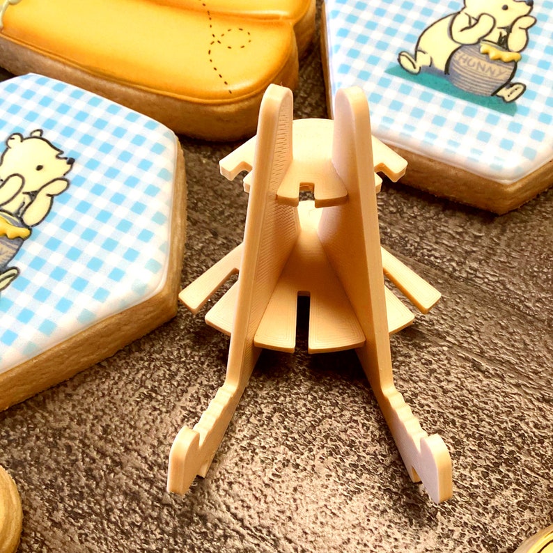 May include: A beige plastic stand with three tiers for displaying cookies or other small treats. The stand has a triangular shape and is designed to hold items securely.