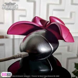 May include: A 3D-printed Cogfly figure from Hollow Knight: Silksong, featuring a metallic silver body and vibrant magenta wings. The figure has a USB cable inserted. The text "Hollow Knight: Silksong" is visible.