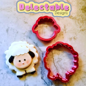 May include: Two red plastic cookie cutters shaped like a sheep. One cutter is a full sheep outline, the other is just the head. A decorated sugar cookie shaped like a sheep is also shown.