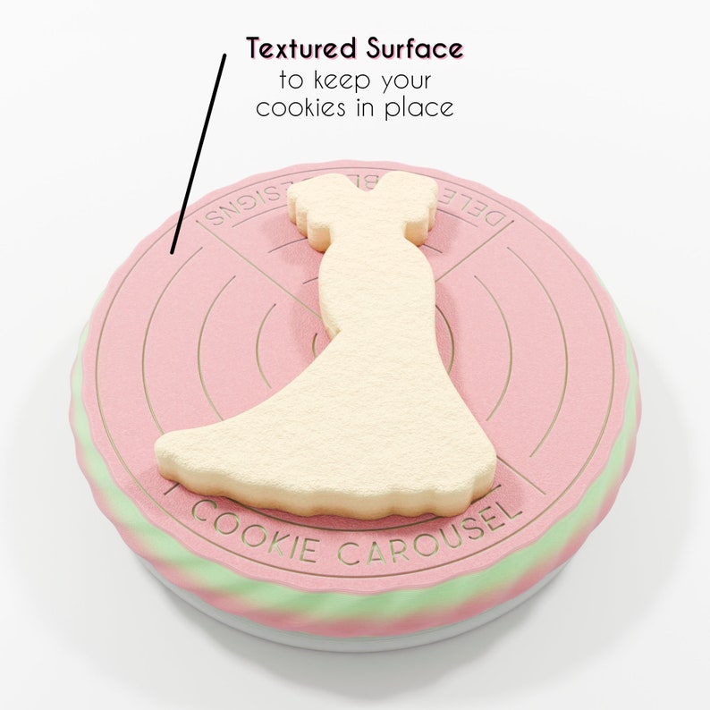 Op de afbeelding: Een roze en groene koekjescarrousel met een getextureerd oppervlak om koekjes op hun plaats te houden. De carrousel heeft de woorden "Cookie Carousel" en "Designs" erop gedrukt. Een witte koek in de vorm van een jurk staat op de carrousel.