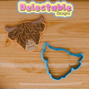 Op de afbeelding: Een bruine en beige koek die is versierd om eruit te zien als een Highland-koeienkop, naast een blauwe plastic koekjesvormer in de vorm van een Highland-koeienkop. De koekjesvormer staat op een houten oppervlak. De tekst "Delectable Designs" is zichtbaar in de rechterbovenhoek van de afbeelding.
