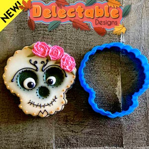 May include: A decorated sugar skull cookie with pink rose accents and a blue skull-shaped cookie cutter. The cookie features black and green details, with the text "Delectable Designs" in the background.