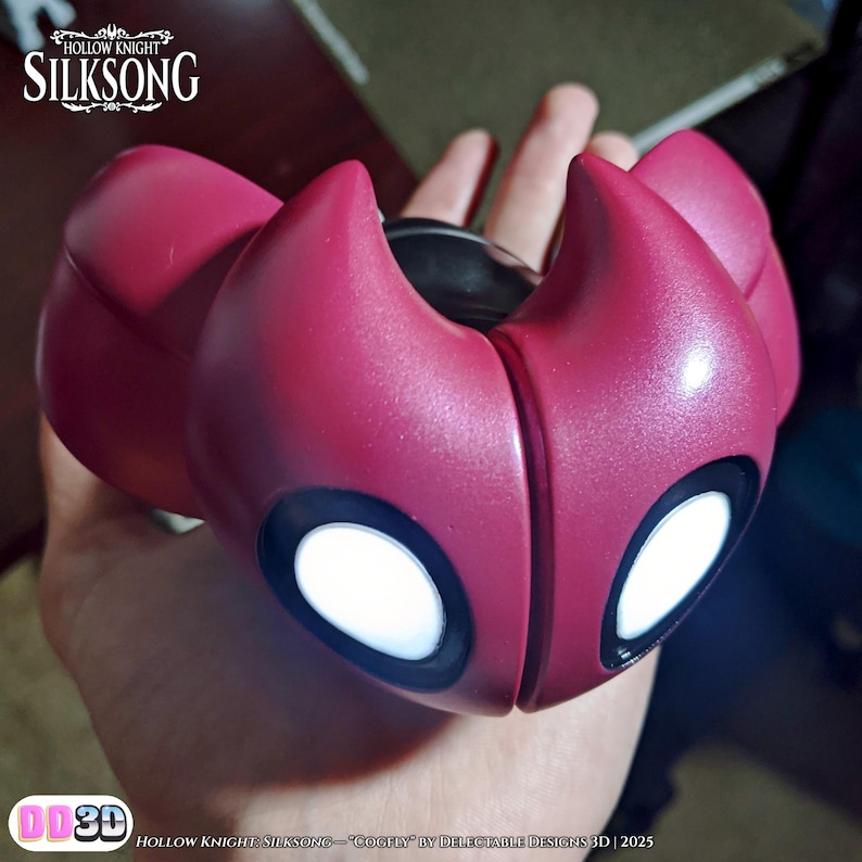 May include: A burgundy-colored mask with white eye-shaped accents and a black center section. The mask is held in a hand. The top left corner of the image has the text "HOLLOW KNIGHT SILKSONG". The bottom left corner has the text "DD3D".