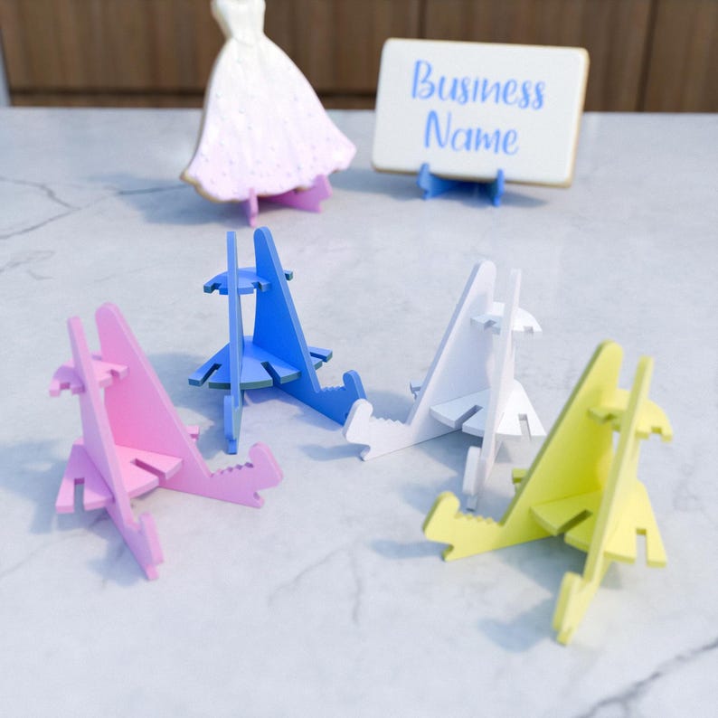 May include: Five phone stands in different colors: pink, blue, white, yellow, and a white stand with the text "Business Name". The stands are made of cardboard and are designed to hold a phone upright.