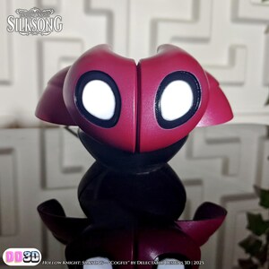 May include: A red and black Cogfly figurine from Hollow Knight: Silksong, with large white eyes and a stylized design. The figurine has a glossy finish and is a 3D printed collectible. The text "HOLLOW KNIGHT SILKSONG" is visible at the top.