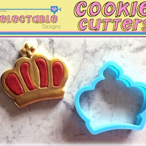 May include: A blue plastic cookie cutter in the shape of a crown. The cutter is on a white marble surface. A gold and red cookie in the shape of a crown is also on the surface.