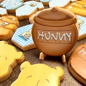 May include: A collection of Winnie the Pooh themed sugar cookies. The cookies are decorated with various images, including Winnie the Pooh, a honey pot, and a bee. The cookies are on a brown surface.