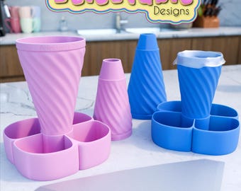May include: Two sets of pink and blue silicone ice cream cone holders. Each set includes a cone holder, a smaller cone holder, and a four-section base. The base has four compartments for holding ice cream scoops. The text "Delectable Designs" is visible at the top of the image.
