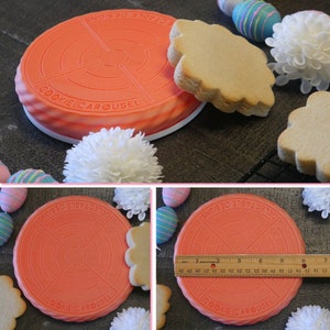 Op de afbeelding: Een rode koekcarrousel met de tekst "Delectable Designs" en "Cookie Carousel" erop. De carrousel wordt gebruikt om koekjes te versieren met verschillende ontwerpen. Er zijn twee koekjes op de carrousel, een is een gewone ronde koek en de andere is een koek met een gegolfde rand.