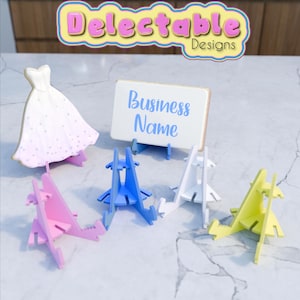 May include: Five colourful cardboard stands for displaying small items such as cookies, cards, or photos. The stands are blue, pink, white, and yellow. A white cookie shaped like a wedding dress is on a stand in the background. A white card with the text "Business Name" is on a stand in the centre.