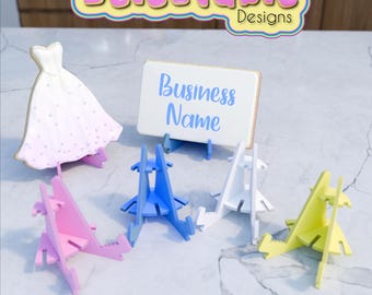 May include: Five colorful cardboard stands for displaying small items like cookies, cards, or photos. The stands are blue, pink, white, and yellow. A white cookie shaped like a wedding dress is on a stand in the background. A white card with the text "Business Name" is on a stand in the center.