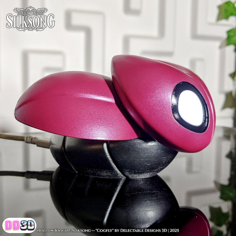 May include: A magenta Cogfly figurine from Hollow Knight: Silksong. The figurine has a rounded body with a black base and a magenta top with a white eye. The text "Hollow Knight Silksong" and "DD3D" are visible.