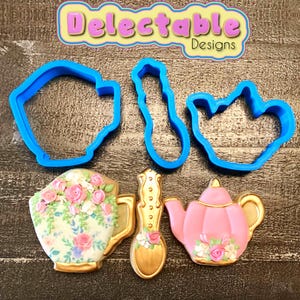 Op de afbeelding: Een assortiment koekjessnijders en versierde koekjes. De blauwe plastic uitsteekvormen zijn gevormd als een theekopje, lepel en theepot. De koekjes zijn versierd met bloemmotieven en gouden accenten. De tekst "Delectable Designs" staat bovenaan.