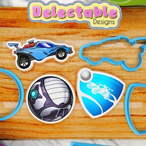 Op de afbeelding: Een set van vijf blauwe koekjesvormen in de vorm van een auto, een bal, een logo met een auto en twee andere vormen. De koekjesvormen liggen op een houten oppervlak met de woorden "Delectable Designs" gedrukt op een bord erboven.