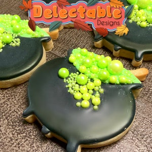 May include: Two black and brown sugar cookies decorated with green icing and green sprinkles. The cookies are shaped like cauldrons. The cookies are decorated with the text "Delectable Designs".