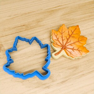 May include: A blue plastic cookie cutter shaped like a maple leaf, next to a decorated sugar cookie in the shape of a maple leaf.