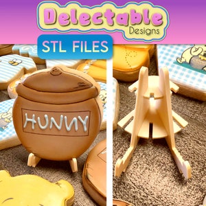 May include: A brown ceramic honey pot cookie with the word "Hunny" written on it. The cookie is on a white stand. The stand is made of plastic and has a simple design. The cookie is part of a set of Winnie the Pooh themed cookies.