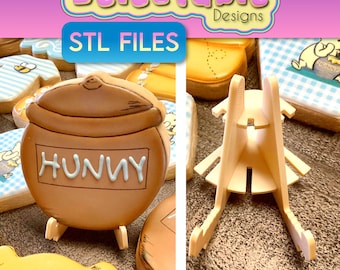 May include: A brown ceramic honey pot cookie with the word "Hunny" written on it. The cookie is on a white stand. The stand is made of plastic and has a simple design. The cookie is part of a set of Winnie the Pooh themed cookies.