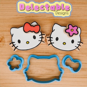 May include: Two white cookie cutters shaped like Hello Kitty faces, one with a red bow and the other with a pink flower. There are also three smaller cookie cutters: a flower, a bow, and a Hello Kitty face.
