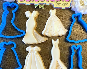 May include: Five cookie cutters in the shape of wedding dresses, with four decorated cookies in the same shapes. The cookie cutters are blue plastic and the cookies are white with pearl-like decorations. The text "Delectable Designs" is in the top center of the image.
