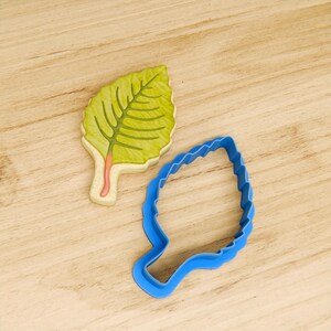 May include: A blue plastic leaf-shaped cookie cutter and a decorated sugar cookie in the shape of a leaf.