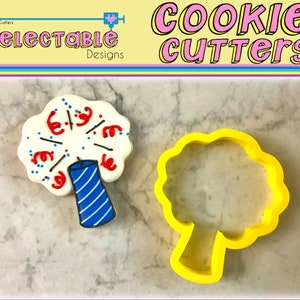 May include: A yellow plastic cookie cutter shaped like a tree with a rounded top and a thick trunk. Next to it is a decorated cookie shaped like a firework with a blue and white striped candle.