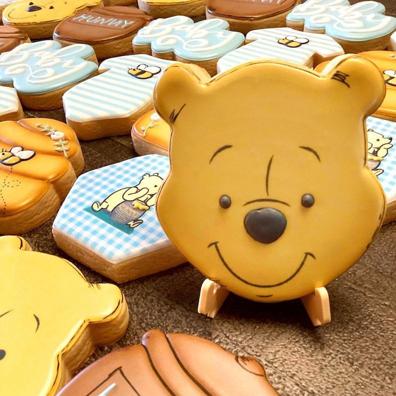 May include: A collection of Winnie the Pooh themed sugar cookies decorated with blue and white icing. The cookies feature Winnie the Pooh's face, a honey pot, and a blue and white checkered pattern.