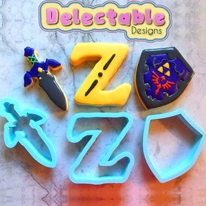 Puede incluir: Un juego de cortadores de galletas con forma de espada, escudo y la letra Z. Los cortadores son azules y están hechos de plástico. Los cortadores están sobre una superficie de mármol blanco.