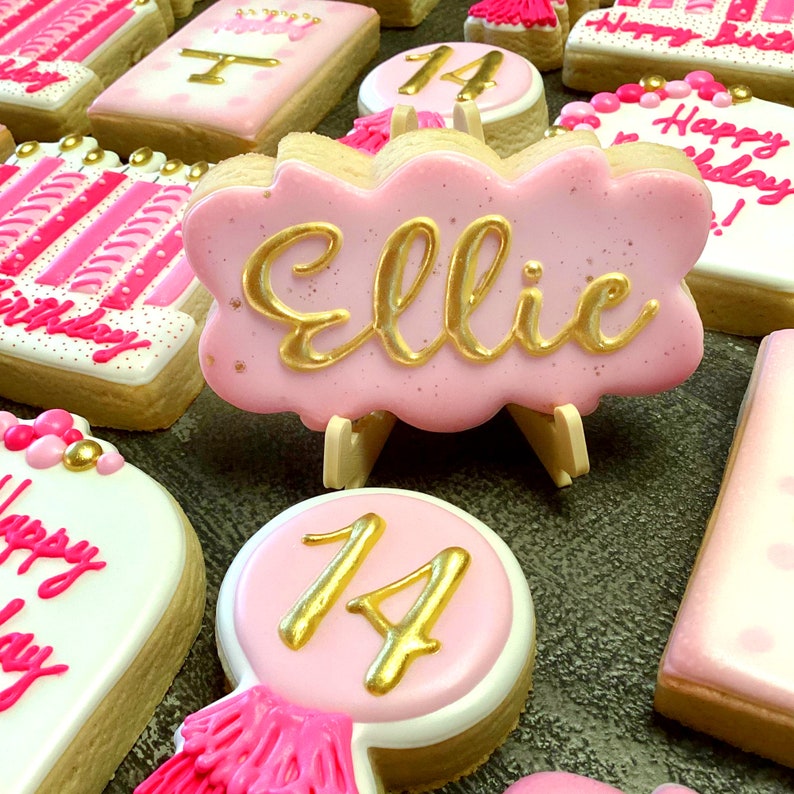 May include: Pink and gold decorated sugar cookies with the name "Ellie" and the number "14" on them. The cookies are decorated with gold writing and pink frosting.