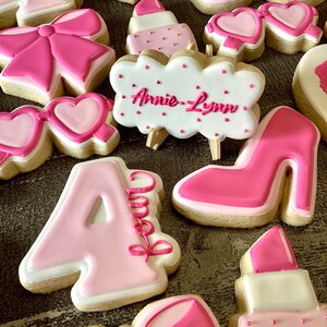 May include: A collection of pink and white decorated sugar cookies with various designs, including a bow, a high heel shoe, a lipstick, sunglasses, and the name "Annie-Lynn".