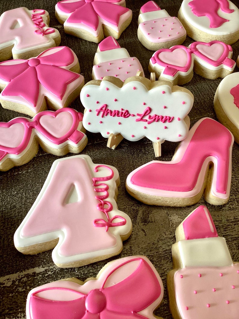 May include: A collection of pink and white decorated sugar cookies with various designs, including a bow, a high heel shoe, a lipstick, sunglasses, and the name "Annie-Lynn".