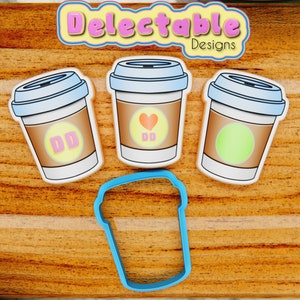 Puede incluir: Tres cortadores de galletas de color blanco con forma de taza de café con diferentes diseños. La primera taza tiene un corazón rosa y las letras DD en su interior. La segunda taza tiene un círculo verde. La tercera taza está en blanco. También se incluye un cortador de galletas azul con forma de taza de café.