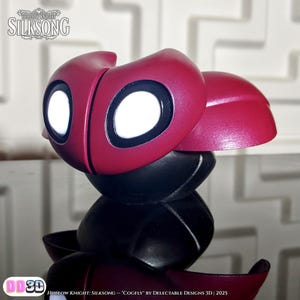 May include: A 3D-printed Cogfly figurine from Hollow Knight: Silksong. The figurine features a burgundy head with large white eyes and a black body. The text "Hollow Knight: Silksong" and "Cogfly" are visible.