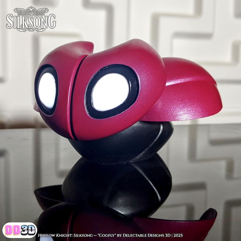 May include: A 3D-printed Cogfly figurine from Hollow Knight: Silksong. The figurine features a burgundy head with large white eyes and a black body. The text "Hollow Knight: Silksong" and "Cogfly" are visible.
