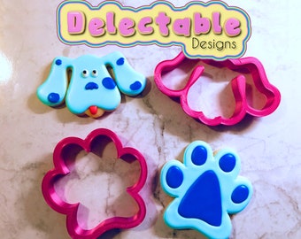 May include: Blue's Clues cookie cutters, featuring a paw print, a paw print cookie, and a blue dog face cookie cutter. The cookie cutters are pink and the cookies are decorated with blue icing.