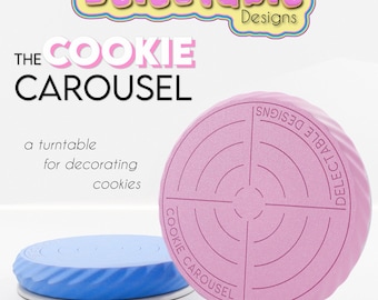 May include: The Cookie Carousel is a pink and blue turntable for decorating cookies. It has a circular design with concentric circles and the text "Delectable Designs" and "Cookie Carousel" printed on it.