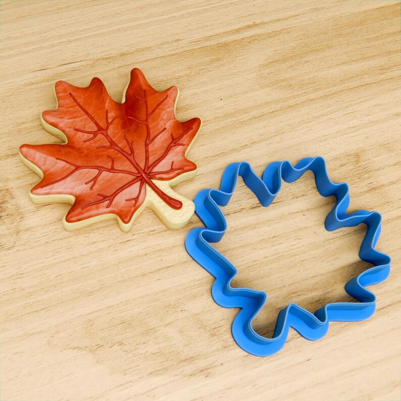 May include: A blue plastic cookie cutter shaped like a maple leaf, next to a decorated maple leaf cookie.