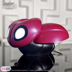 May include: A 3D-printed Cogfly figurine from Hollow Knight: Silksong. The figurine features a maroon head with large white eyes outlined in black, atop a black body. The text "HOLLOW KNIGHT SILKSONG" is visible. The text "COGFLY" is also visible.