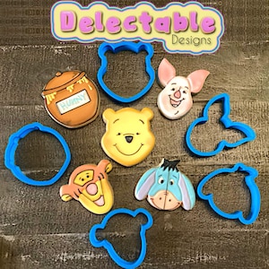 May include: A collection of Winnie the Pooh character-themed cookies and blue cookie cutters. The cookies include Pooh Bear, Piglet, Tigger, Eeyore, and a honey pot. The text "Delectable Designs" is in the top left corner.