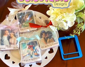 May include: Four decorated cookies with personalized photos and messages. The cookies are packaged in clear cellophane bags with ribbons. The cookies are on a white plate with heart-shaped cutouts. A blue plastic cookie cutter in the shape of a Polaroid photo is on a wooden surface.