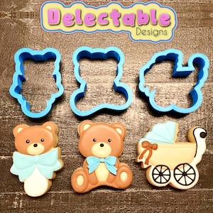 Può includere: Un set di tre tagliapasta in plastica blu a forma di orsacchiotto, orsacchiotto seduto e passeggino. I tagliapasta sono su una superficie di legno accanto a tre biscotti decorati. I biscotti sono decorati con glassa blu e un fiocco. Il testo "Delectable Designs" è visibile nella parte superiore centrale dell'immagine.