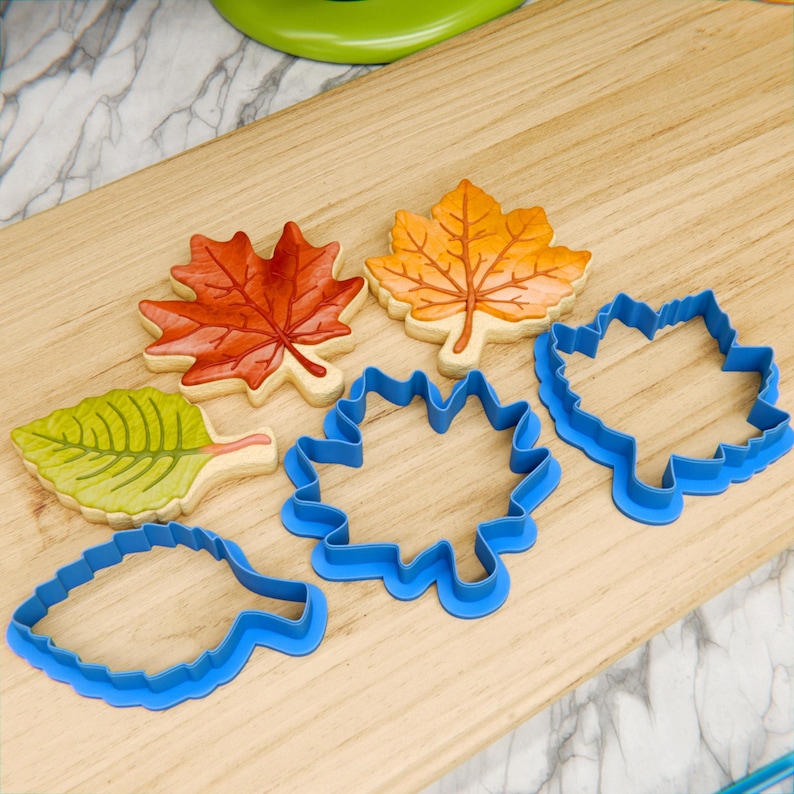 May include: Five cookie cutters in the shape of leaves. Four of the cutters are blue plastic and one is a brown, orange, and yellow leaf cookie cutter.