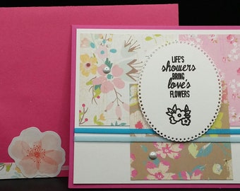 May include: A handmade greeting card with a pink envelope. The card features floral patterned paper, a white oval with the text "Life's showers bring love's flowers," and a small flower illustration. Embellished with small silver gems.
