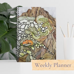 May include: A weekly planner with a spiral binding and a colorful cover featuring a stylized illustration of a fox sleeping in a cave-like formation with a brown, green, and white color scheme. The text "Weekly Planner" is printed on a wooden tag.