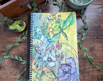 May include: A spiral bound journal with a colorful watercolor illustration of flowers, birds, and leaves. The cover features a yellow flower, blue birds, and green leaves on a yellow background.